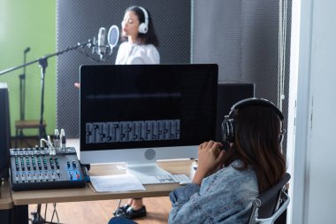 Genç Asyalı kadın vokalist kulaklık takarak profesyonel bir stüdyoda mikrofon önünde şarkı kaydettiği için Software 'ın bilgisayar monitörü kadın teknisyeni.