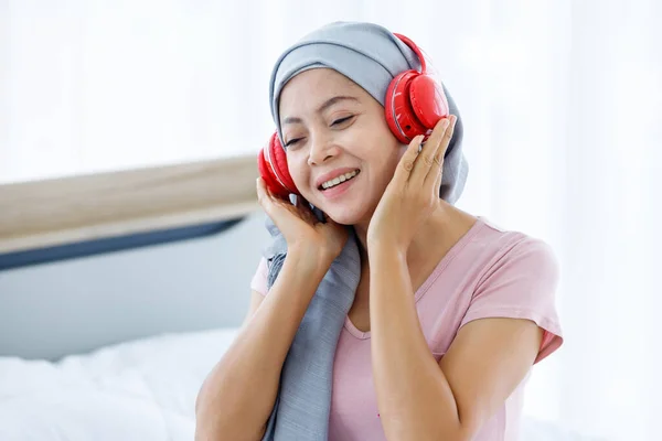 Happy a asian women disease mammary cancer patient with pink ribbon wearing headscarf in headphones is listening to music After treatment to chemotherapy sit on bed In the bedroom at the house
