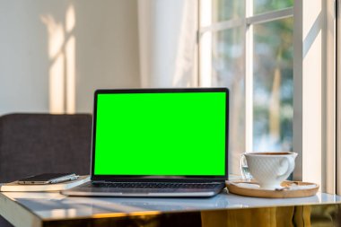 Kahve dükkanının arkasındaki pencerenin yanındaki masada kahve fincanı ve akıllı telefonu olan boş ekranlı dizüstü bilgisayarın bir kopyası, yeşil ekran.