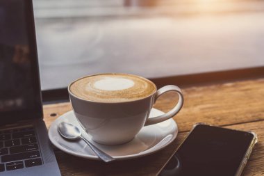 Kahve fincanında sütlü sütlü sütlü sütlü sütlü sütlü sütlü latte ve iş konsepti sırasında kafedeki ahşap ofis masasında akıllı telefon.