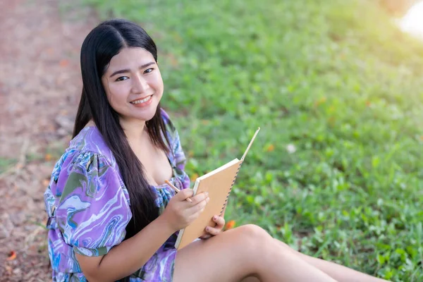 Happy Relaxing Portrait of freelancer asian woman Wear purple dress while working holding diary book writing note while sitting on green grass lawn beside a reservoir at the city park outdoors.