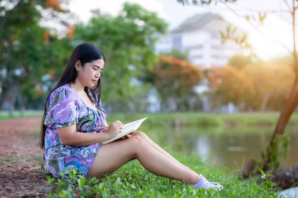 Happy Relaxing Portrait of freelancer asian woman Wear purple dress while working holding diary book writing note while sitting on green grass lawn beside a reservoir at the city park outdoors.