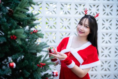 Kırmızı Noel Baba 'lı şirin gülümseyen Asyalı kız Noel ve yeni yıl boyunca Noel ağacı arkaplan dekorasyonunda fotoğraf kutlaması kutlaması yaparken kameraya bakıyor..