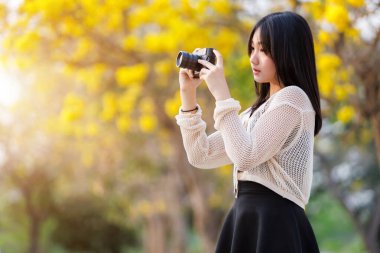 Çekici genç kızın Asyalı gülümsemesi. Sarı Tabebuia Chrysotricha çiçeklerinin içinde elinde bir kamera. Baharın ilk günü Tayland 'da akşam arkaplanında parkla birlikte..