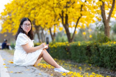 Asyalı Mutlu Rahatlatıcı Portre Kadını. Sarı Tabebuia Chrysotricha Çiçekleri 'nde elinde tuttuğu günlük defteri. Bahar günü Tayland' da akşam arkaplanı parkı..