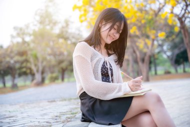 Çekici bir genç kızın portresi Asyalı gülümsüyor. Sarı Tabebuia Chrysotricha çiçeklerinde elinde bir günlük defteri tutuyor. Bahar günü Tayland 'da akşam arkaplanında parkla birlikte..
