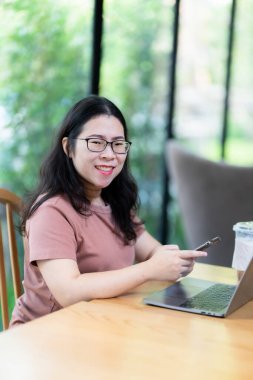 Asyalı serbest çalışanlardan memnun olan iş kadını akıllı telefondan yazı okuyor. Kafede dizüstü bilgisayar ve kahve fincanı ile çalışıyor. İş hayatı tarzı iletişim kavramı.