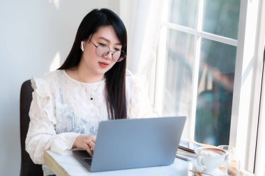 Asyalı serbest çalışanlardan memnun. İş kadını. Kablosuz kulaklık takıyor. Laptop bilgisayarıyla çalışıyor.