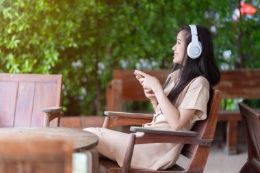 Beyaz kablosuz kulaklık takan, müzik dinleyen ve güneşli bir günde parkta ya da kafede kullanan Asyalı genç bir kız.