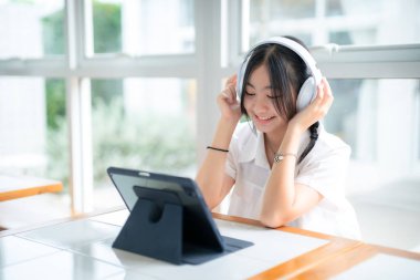 Şirin gülümsemeler Asyalı çekici küçük sevimli kız kulaklık takıyor ve müzik dinliyor kafede çalışan dijital tabletleri kullanıyor. Çevrimiçi eğitim, öğrenim konsepti..