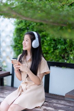 Beyaz kablosuz kulaklık takan, müzik dinleyen ve güneşli bir günde parkta ya da kafede kullanan Asyalı genç bir kız.