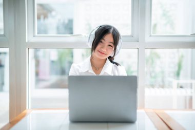 Şirin gülüşler Asyalı, kulaklık takan, müzik dinleyen, kulaklık takan, kafede çalışan bilgisayarlı çekici bir kız. Çevrimiçi eğitim, gelişim konsepti..