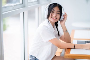 Şirin gülüşler Asyalı, kulaklık takan, müzik dinleyen, kulaklık takan, kafede çalışan bilgisayarlı çekici bir kız. Çevrimiçi eğitim, gelişim konsepti..