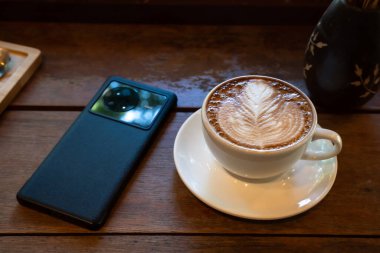 Sütlü sütlü sütlü sütlü sıcak kahve fincanı, ahşap masanın üstünde akıllı telefon. Kafedeki bir kafede kahvaltı olarak, iş konsepti sırasında, klasik tarzda.