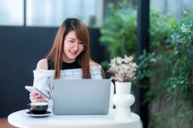 Asyalı serbest çalışan iş kadını bilgisayarla internet, sohbet, blog ve akıllı telefon, kafede defter gibi şeyleri taramak için günlük olarak bilgisayarla çalışarak akıllı telefondan mesaj okuyor..