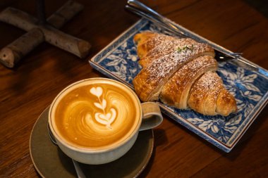 Sütlü sütlü sütlü sütlü sütlü sıcak kahve ve ahşap masa üzerinde kruvasan. Kafedeki bir kafede kahvaltı olarak, iş konsepti sırasında, klasik tarzda.
