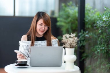 Asyalı serbest çalışan iş kadını bilgisayarla internet, sohbet, blog ve akıllı telefon, kafede defter gibi şeyleri taramak için günlük olarak bilgisayarla çalışarak akıllı telefondan mesaj okuyor..