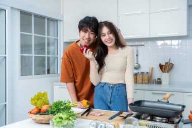 Sevgi dolu genç bir çiftin mutlu portresi. Asyalı neşeli bir şekilde yemek pişirmeyi seviyor. Sebze, et, ekmek pişiriyor.