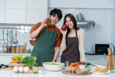 Sevgi dolu genç bir çiftin mutlu portresi. Asyalı neşeli bir şekilde yemek pişirmeyi seviyor. Sebze, et, ekmek pişiriyor.
