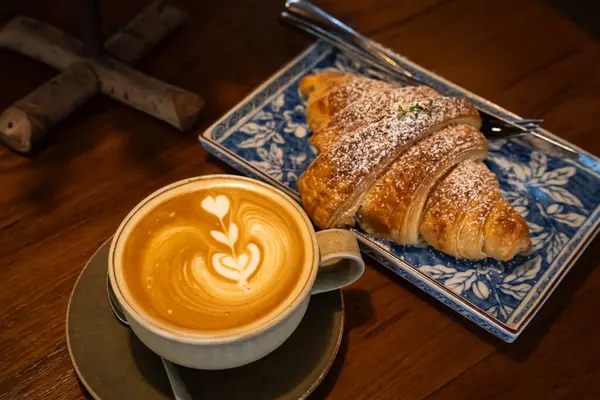 Sütlü sütlü sütlü sütlü sütlü sıcak kahve ve ahşap masa üzerinde kruvasan. Kafedeki bir kafede kahvaltı olarak, iş konsepti sırasında, klasik tarzda.