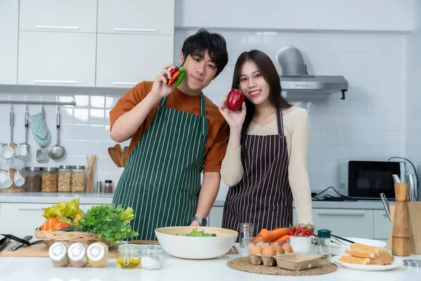 Sevgi dolu genç bir çiftin mutlu portresi. Asyalı neşeli bir şekilde yemek pişirmeyi seviyor. Sebze, et, ekmek pişiriyor.