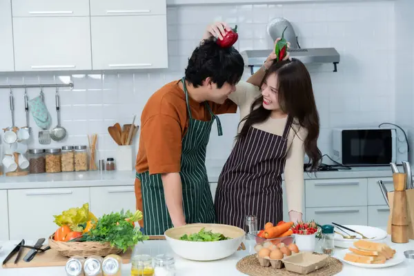 Sevgi dolu genç bir çiftin mutlu portresi. Asyalı neşeli bir şekilde yemek pişirmeyi seviyor. Sebze, et, ekmek pişiriyor.