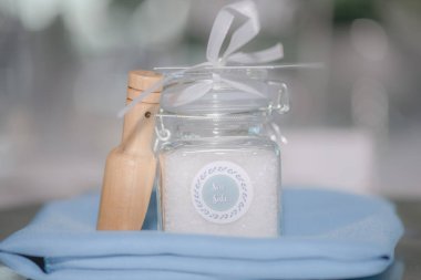 Beautiful Sea Salt filled clear jar with white bow wedding favor with wood on blue blanket in front of white bokeh clipart