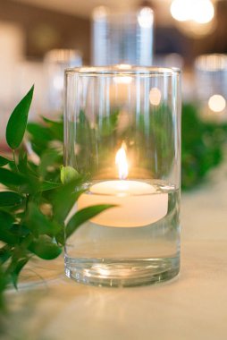 Uzun cam vazo, içinde yanan çay ışığı mumu olan yeşil bitkilerle düğün masasında yüzen.