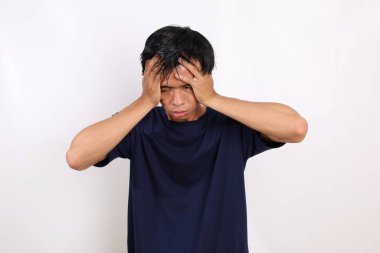Stresli Asyalı genç adam başını tutarken ayakta duruyor. Beyaz arkaplanda izole