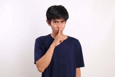 Genç Asyalı bir adam, beyaz arka planda dudaklarını tutarken ayakta duruyor.