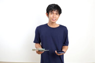 Endonezya banknotları ve cep telefonu elindeyken ayakta duran mutlu Asyalı genç adam. Beyazda izole edilmiş