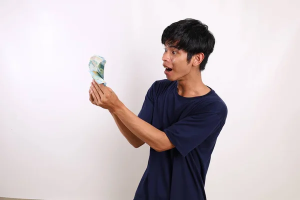 令人惊讶的年轻亚洲男子站在那里 手里拿着印度尼西亚钞票 与白种人隔离 — 图库照片