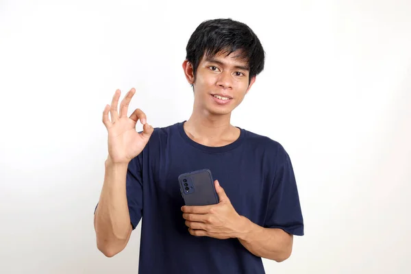 快乐的年轻亚洲男人拿着手机站在那里 摆出一副不错的手势 与白种人隔离 — 图库照片