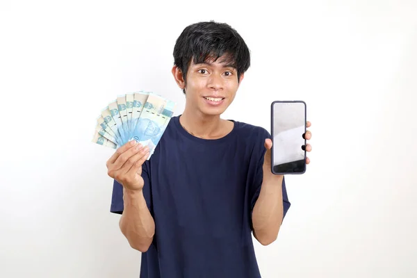 快乐的亚洲年轻人站在那里 手里拿着印尼钞票 展示着空白的手机屏幕 与白种人隔离 — 图库照片