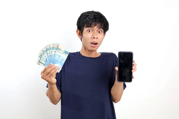 令人惊讶的是 年轻的亚洲男子拿着印尼钞票站在那里 展示空白的手机屏幕 与白种人隔离 — 图库照片