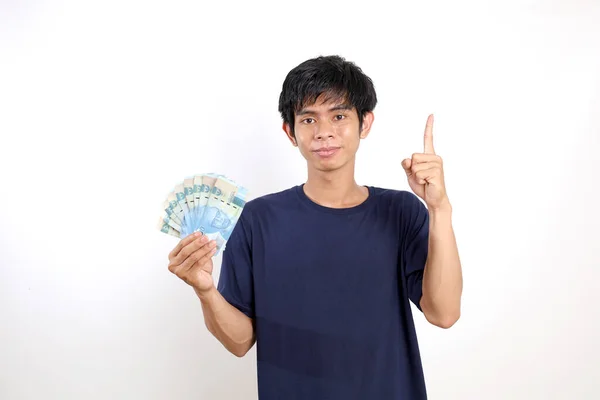 快乐的年轻亚洲男人站在那里 手里拿着印尼钞票 一只手指头 或者指着上面 — 图库照片