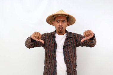 Mutsuz Asyalı çiftçi baş parmağını kaldırırken ayakta duruyor. Beyaz arkaplanda izole