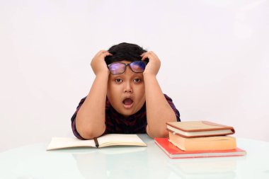 Yorgun Asyalı okul çocuğu ödevini yaparken oturuyor. Beyaz arkaplanda izole