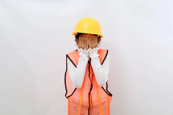 Stressed Asian little girl in the construction helmet as an engineer standing while covering her face. Isolated on white