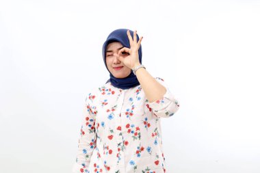 Cheerful asian muslim woman standing while making okay hand gesture near her eye. Isolated on white