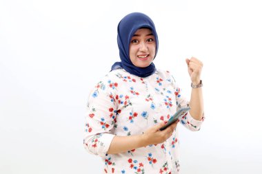 Excited asian muslim woman standing while holding a cell phone and showing fist hand.