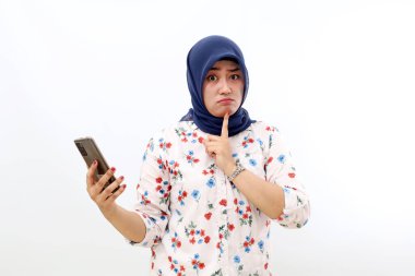 Thoughtful asian muslim woman holding a cell phone while thinking something. Isolated on white