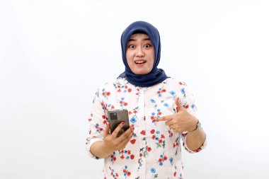 Surprised asian muslim woman holding a cell phone while pointing on it. Isolated on white background