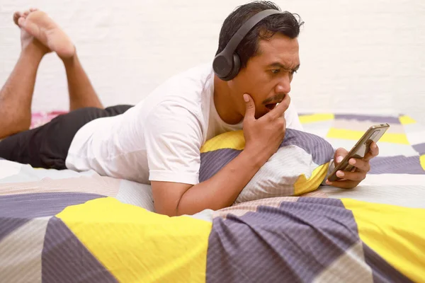 Shocked and angry asian man holding a cell phone and using headphone on the bed at home