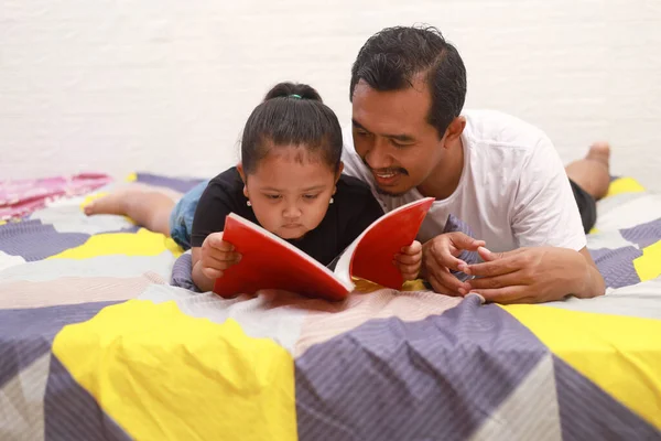 Asian dad accompanies his daughter reading a book on the bed at home