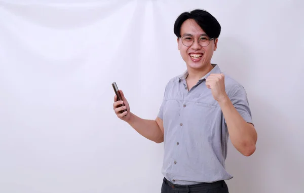 携帯電話を持ちながらカメラを見ながら立っている幸せな興奮したアジア人男性 白に隔離された — ストック写真