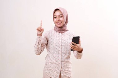 Gülümseyen Asyalı müslüman kadın parmağını gösterirken cep telefonu tutuyor. Beyazda izole edilmiş