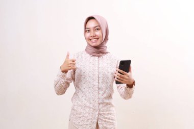 Mutlu Asyalı genç Müslüman kız parmağını kaldırırken elinde cep telefonu tutuyor. Beyazda izole edilmiş
