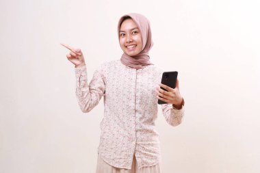 Yan tarafı gösterirken cep telefonu tutan heyecanlı Asyalı genç bir müslüman kız. Beyaz arkaplanda izole