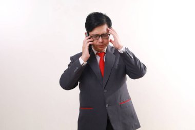 Stresli Asyalı iş adamı telefonda konuşurken başını tutuyor. Beyazda izole edilmiş